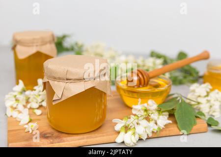 Süßes Honigglas umgab die Akazienblüten des Frühlings. Honig fließt aus einem Löffel in ein Glas. Gläser von klarem frischen Akazienhonig auf Holzgrund. Stockfoto