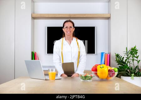 Porträt einer lächelnden jungen Ärztin, Ernährungswissenschaftlerin, die in einem hellen, modernen Bürozimmer mit Waage und Maßband steht Stockfoto