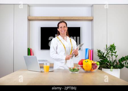 Porträt einer lächelnden jungen Ärztin, Ernährungswissenschaftlerin, die in einem hellen, modernen Bürozimmer mit Waage und Maßband steht Stockfoto
