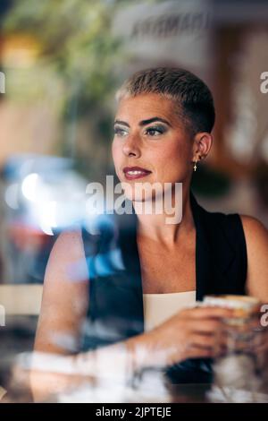 Die Erwachsene Frau schaut selbstbewusst aus dem Fenster, während sie Kaffee trinkt, Blick von außen, Porträt Stockfoto