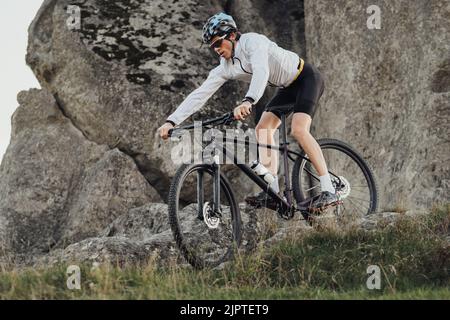 Ausgerüsteter Profi-Radler fährt mit seinem Mountainbike eine Steigung hinunter, Sportler fährt auf dem Fahrrad vom Berg herunter, Riesenstein auf dem Hintergrund Stockfoto