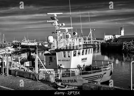 Ein Wachboot im Hafen, das mit Antennen und Radar bristillt, wird in einem Abendlicht gefangen. Ein Leuchtturm ist dahinter und ein Himmel mit Wolken ist oben. Stockfoto