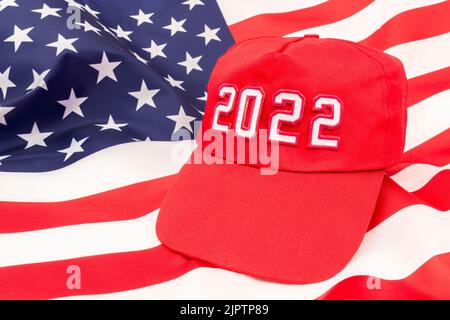 Red MAGA-Typ Hut mit 2022 auf Hintergrund US Stars and Stripes Flagge. Für republikanische Siege bei den Midterm-Wahlen in den USA im November 2022 und der republikanischen Roten Welle. Stockfoto