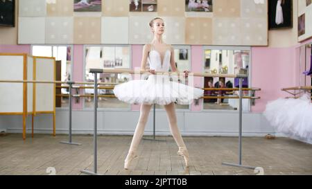 Im Ballettsaal ist das Mädchen im weißen Pack beim Ballett verlobt, probt Roleve, geht auf die Zehen, in Spitzenschuhen, Junge Ballerina steht am Geländer im Ballettsaal. Hochwertige Fotos Stockfoto