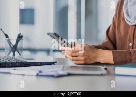 Nahaufnahme des Fotos selektiver Fokus auf die Hände von Geschäftsfrauen, die am Telefon tippen, Frau, die an einem Tisch mit Dokumenten und einem Laptop in einem modernen Büro arbeitet Stockfoto