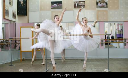 Im Ballettsaal werden Mädchen in weißen Ballettröcken beim Ballett verlobt, üben Haltung, Junge Ballerinas stehen in Spitzenschuhen auf den Zehen, heben die Beine elegant nach hinten, beim Geländer im Ballettsaal. Hochwertige Fotos Stockfoto