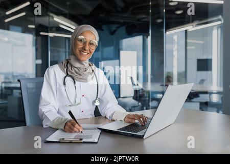 Porträt einer Ärztin in Hijab, muslim, der in einem modernen Büro in der Klinik arbeitet, lächelt und die Kamera anschaut, Dokumente ausfüllt, Laptop benutzt Stockfoto