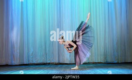 Ballettprobe, auf der Bühne des alten Theatersaals. Die junge Ballerina in lila schwarzem Kleid und Spitzenschuhen tanzt elegant bestimmte Ballettbewegungen, hebt ihr Bein nach hinten hoch. Hochwertige Fotos Stockfoto