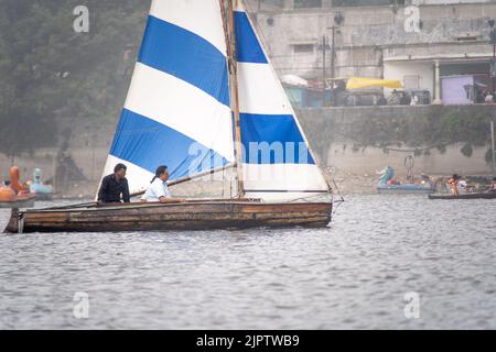 Kleines buntes Segelboot mit Leuten, die darauf sitzen, vorbei an Ruderbooten und Tretbooten an einem nebligen Tag mit Gebäuden im Hintergrund auf dem See von Stockfoto