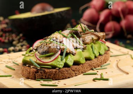 Ein Avocado-Toast mit Pilzen, Rettich, Sesam und Schnittlauch auf einem Holzschneidebrett. Stockfoto