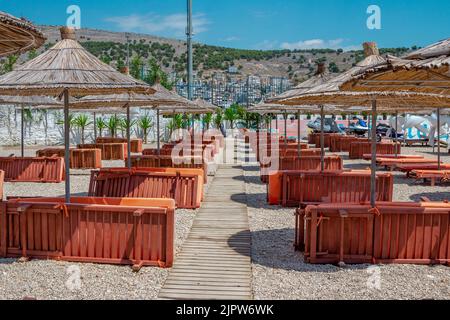 Saranda, Albanien - 5. August 2020: Blick auf den leeren Strand - Sonnenliegen und Strohschirmschirme in der Nähe von Meerwasser, Stadtgebäuden und Bergen am Horizont. Stockfoto