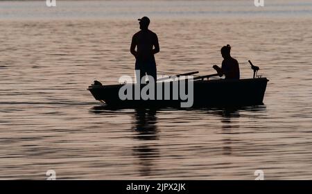 Eutin, Deutschland. 16. August 2022. 16.08.2022, Eutin. Zwei junge Männer fischen von einem Boot am Kellersee im Hintergrund der Abendsonne. Der eine (l) verwendet eine Spinnstange, der andere eine Fliegenstange. Die beiden Angler und das Fischerboot sind nur als Silhouetten sichtbar, wie eine Silhouette. Quelle: Wolfram Steinberg/dpa Quelle: Wolfram Steinberg/dpa/Alamy Live News Stockfoto