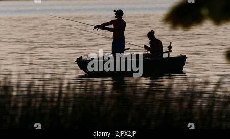 Eutin, Deutschland. 16. August 2022. 16.08.2022, Eutin. Zwei junge Männer fischen von einem Boot am Kellersee im Hintergrund der Abendsonne. Der eine (l) verwendet eine Spinnstange, der andere eine Fliegenstange. Die beiden Angler und das Fischerboot sind nur als Silhouetten sichtbar, wie eine Silhouette. Quelle: Wolfram Steinberg/dpa Quelle: Wolfram Steinberg/dpa/Alamy Live News Stockfoto