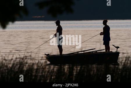 Eutin, Deutschland. 16. August 2022. 16.08.2022, Eutin. Zwei junge Männer fischen von einem Boot am Kellersee im Hintergrund der Abendsonne. Der eine (l) verwendet eine Spinnstange, der andere eine Fliegenstange. Die beiden Angler und das Fischerboot sind nur als Silhouetten sichtbar, wie eine Silhouette. Quelle: Wolfram Steinberg/dpa Quelle: Wolfram Steinberg/dpa/Alamy Live News Stockfoto