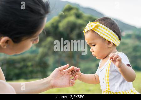 Schöne braune Latina-Mädchen lernen zu gehen, halten ihre Mutter die Hand, in einem offenen Grasfeld an einem schönen Sommertag. Familie concep Stockfoto