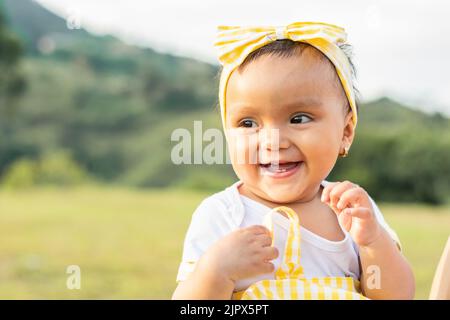 Schönes braunhäutiges Latina-Baby auf einem Außenfeld an einem Sommertag, lächelnd und zeigt ihre kleinen Zähne, die immer noch aus ihrem Zahnfleisch kommen. c Stockfoto