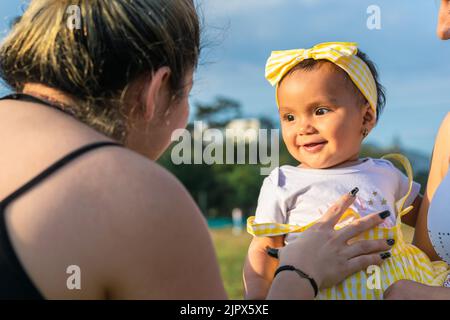 Schönes Baby mit brauner Haut, in den Armen ihrer Mutter, schaut auf ihre Tante, während sie ihren Bauch kitzelt, um ihre neuen Babyzähne zu zeigen. Schöner Sommertag Stockfoto