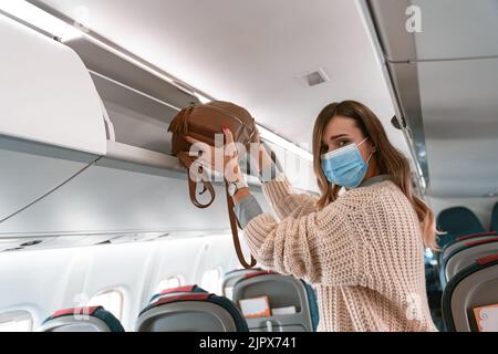 Frau in Maske, die Gepäck während des Boarding in den Gepäckraum im Flugzeug legt Stockfoto