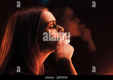 Tausendjährige cool hübsches Mädchen mit ungewöhnlichen gefärbt Frisur rauchen Zigarette in der Nähe von glühenden neon Wand in der Nacht. Blaue Haare, goldenen Pailletten wie Sommersprossen, Nase Stockfoto