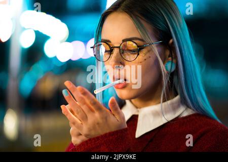 Millennial hübsches Mädchen mit ungewöhnlich gefärbten Frisur Rauchen Zigarette in Vergnügungspark in der Nacht. Blaue Haare, goldene Pailletten als Sommersprossen, Nasenpiercing Stockfoto