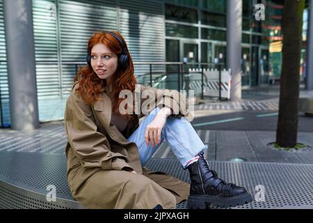 Lächelndes Mädchen mit rothaarigen Hipstern, das Kopfhörer trägt und Musik in der Stadt hört. Stockfoto