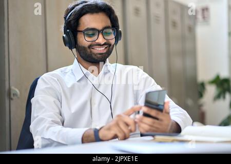 Lächelnder indischer Geschäftsmann, der Kopfhörer trägt und das Smartphone bei der Arbeit benutzt. Stockfoto