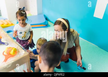 Frischhaltefolie Malerei für die Verbesserung der Kinder Phantasie und Gehirnentwicklung. Kleinkinder und ihre Lehrerin malen mit Bürsten, Rollen und Farben auf einer Frischhaltefolie, die um den hölzernen Regalständer gewickelt ist. Lustige Aktivität für Kinder feine und grobe Motorik im Kindergarten. Hochwertige Fotos Stockfoto