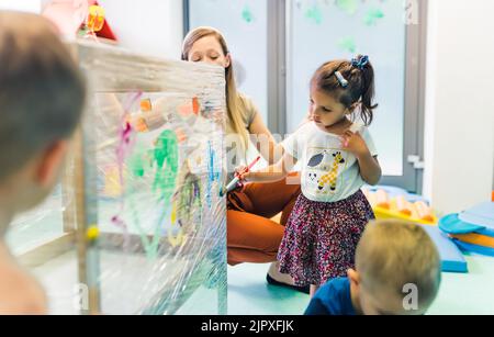 Frischhaltefolie für die Verbesserung der Phantasie von Kindern. Kleinkinder malen mit, Bürsten, Rollen und Farben auf einer Frischhaltefolie, die um den hölzernen Regalständer gewickelt ist. Lustige Aktivität für Kinder Sinnesfertigkeiten, Kreativität, räumliche Aufmerksamkeit und visuelle Wahrnehmungsentwicklung im Kindergarten. Stockfoto