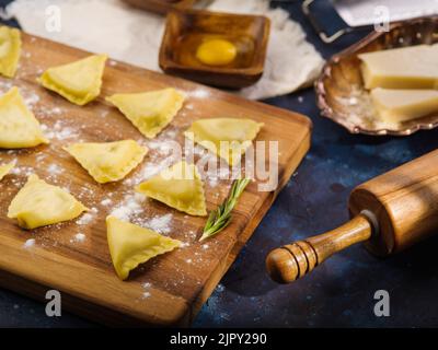 Appetitliche hausgemachte Ravioli auf einem Holzschneidebrett, Zutaten und Küchenutensilien auf blauem Hintergrund. Rezepte der italienischen Küche. Hausmannskost Stockfoto