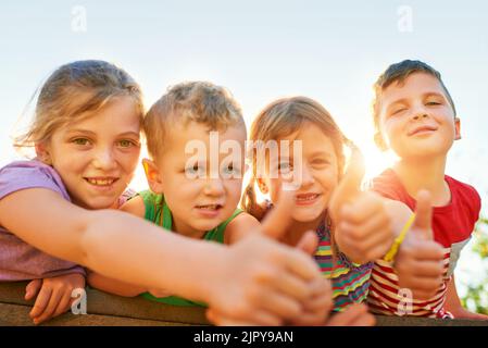 Spielzeit ist die beste Zeit. Porträt einer Gruppe von kleinen Kindern, die Daumen hoch zeigen, während sie im Freien zusammen spielen. Stockfoto