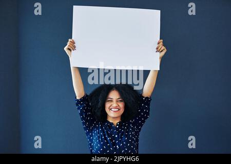 Sagen Sie es laut, sagen Sie es stolz. Studioaufnahme einer jungen Frau, die ein leeres Plakat vor blauem Hintergrund hochhält. Stockfoto