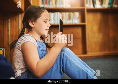 Ich denke, es ist an der Zeit, eine Brille zu bekommen. Ein fokussiertes junges Mädchen, das tagsüber auf einem digitalen Tablet surft, während es in einer Bibliothek sitzt. Stockfoto