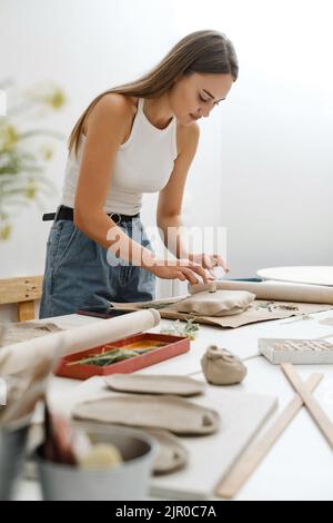 Frau, die Ton rollt, macht keramische Platte im Studio mit Blumenmuster. Handgefertigte kreative Arbeiten. Töpferwerkstatt für Erwachsene. Stockfoto