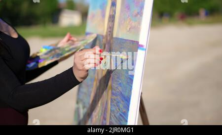 Sommer, im Freien, Nahaufnahme der weiblichen Hände der Künstlerin. Sie trägt Farbe auf die Leinwand mit einem speziellen kleinen Spachtel, mit einer speziellen Zeichentechnik. Hält eine Palette mit Farben. Hochwertige Fotos Stockfoto