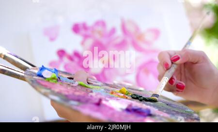 Nahaufnahme, weibliche Hände, Malerin, Künstlerin malt ein Blumenbild, sie hält Palette mit Farben und Pinsel, mischt Farben mit einem Pinsel auf der Palette. Hochwertige Fotos Stockfoto