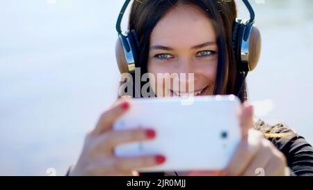 Sommer, im Freien, Porträt einer schönen Frau, mit langen dunklen Haaren, Musik hören auf Kopfhörer von einem Mobiltelefon, Gerät, Video per Telefon zu schießen, macht Selfi. Hochwertige Fotos Stockfoto