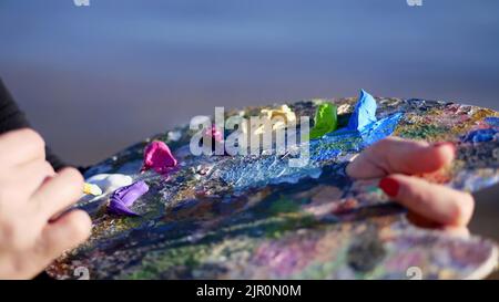 Sommer, im Freien, Nahaufnahme der weiblichen Hände der Künstlerin und eine Palette mit Farben, mischt die Künstlerin Farben mit einem Pinsel auf der Palette. Hochwertige Fotos Stockfoto