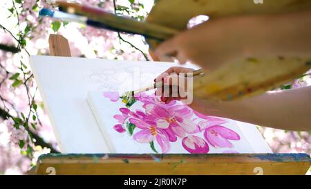 Nahaufnahme, Malerin zeichnen ein Bild von Blumen in blühenden Apfelplantagen, sie malt mit Pinsel, hält Palette mit Farben. Wind fegt blühende Äste über die Staffelei. Hochwertige Fotos Stockfoto