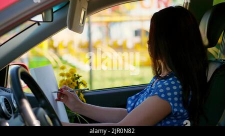 Künstler, schöne Brünette Frau, sitzt im Auto, zeichnet Bleistift Skizze, Zeichnung Strauß von gelben Dandelionen. Zeichnung aus der Natur, der kreative Prozess des Zeichnens. Sommer. Hochwertige Fotos Stockfoto