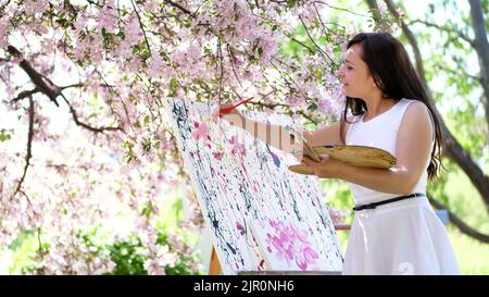 Eine schöne Malerin in weißem Kleid, Künstlerin malt ein Bild von Blumen in blühenden Frühling Apfelgarten, hält sie eine Palette mit Farben und einem Pinsel in den Händen. Hochwertige Fotos Stockfoto
