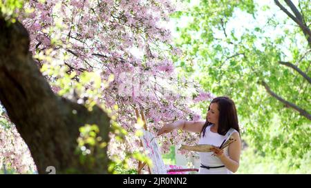 Eine schöne Malerin in weißem Kleid, Künstler malt ein Bild von Blumen in blühenden Frühling Apfelgarten, sie trägt Farben auf die Leinwand mit einem speziellen kleinen Spatel, mit einer speziellen Zeichentechnik. Hält eine Palette mit Farben. Hochwertige Fotos Stockfoto