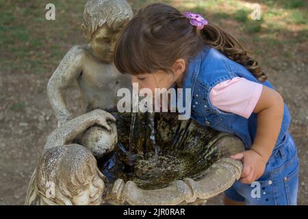 Bild eines entzückenden kleinen Mädchens, das Wasser aus einem geschmückten Brunnen im öffentlichen Park trinkt. Spiele und Outdoor-Leben mit den Kindern während des Urlaubs Stockfoto