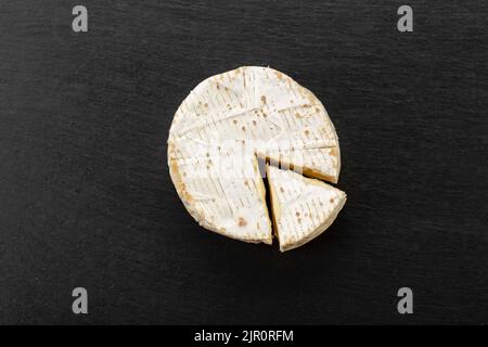 Brie-Käsesorte. Camembert-Käse. Frischer Brie-Käse und eine Scheibe auf einem schwarzen Brett. Italienischer, französischer Käse. Draufsicht. Stockfoto