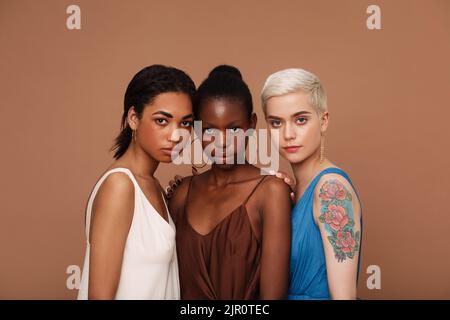 Eine Gruppe von drei Frauen verschiedener Rassen, die vor braunem Hintergrund zusammenstehen Stockfoto