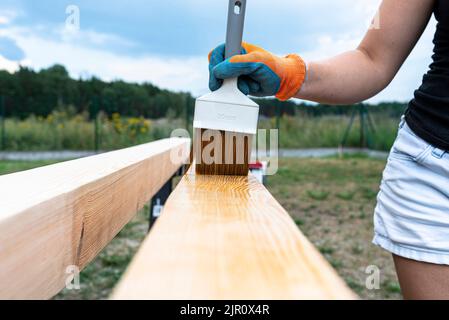 Die Frau imprägniert roh gehobelte Kiefernbalken mit Abmessungen von 100x100mm mit einer farblos imprägnierten. Stockfoto