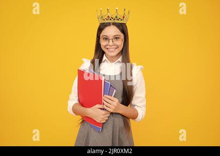 Schulmädchen in Schuluniform und Krone feiern den Sieg auf gelbem Hintergrund. Schulkind hält Bücher. Bildungsabschluss, Sieg und Erfolg. Stockfoto