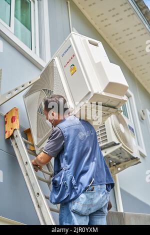 Ein Mann trägt einen schweren Klimaanlagenkompressor auf seiner Schulter, während er eine Leiter klettert. Stockfoto