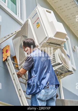 Ein Mann trägt einen schweren Klimaanlagenkompressor auf seiner Schulter, während er eine Leiter klettert. Stockfoto