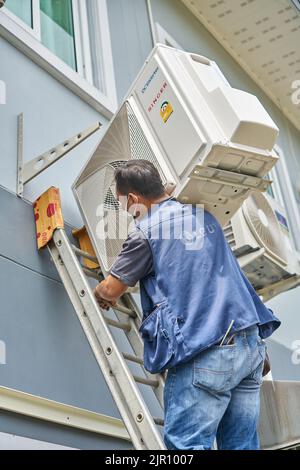 Ein Mann trägt einen schweren Klimaanlagenkompressor auf seiner Schulter, während er eine Leiter klettert. Stockfoto