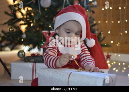 Ein einjähriges Baby öffnet ein Weihnachtsgeschenk. Baby als weihnachtsmann-Eröffnungsgeschenk gekleidet. Süßes Baby neben dem weihnachtsbaum. Begeistert, das Geschenk zu öffnen. Stockfoto